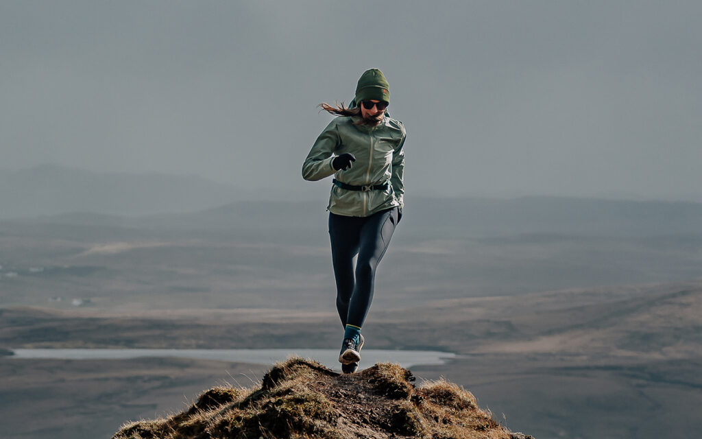 Person in a green jacket and beanie jumps on a rocky hilltop with rolling hills and a distant valley in the background.